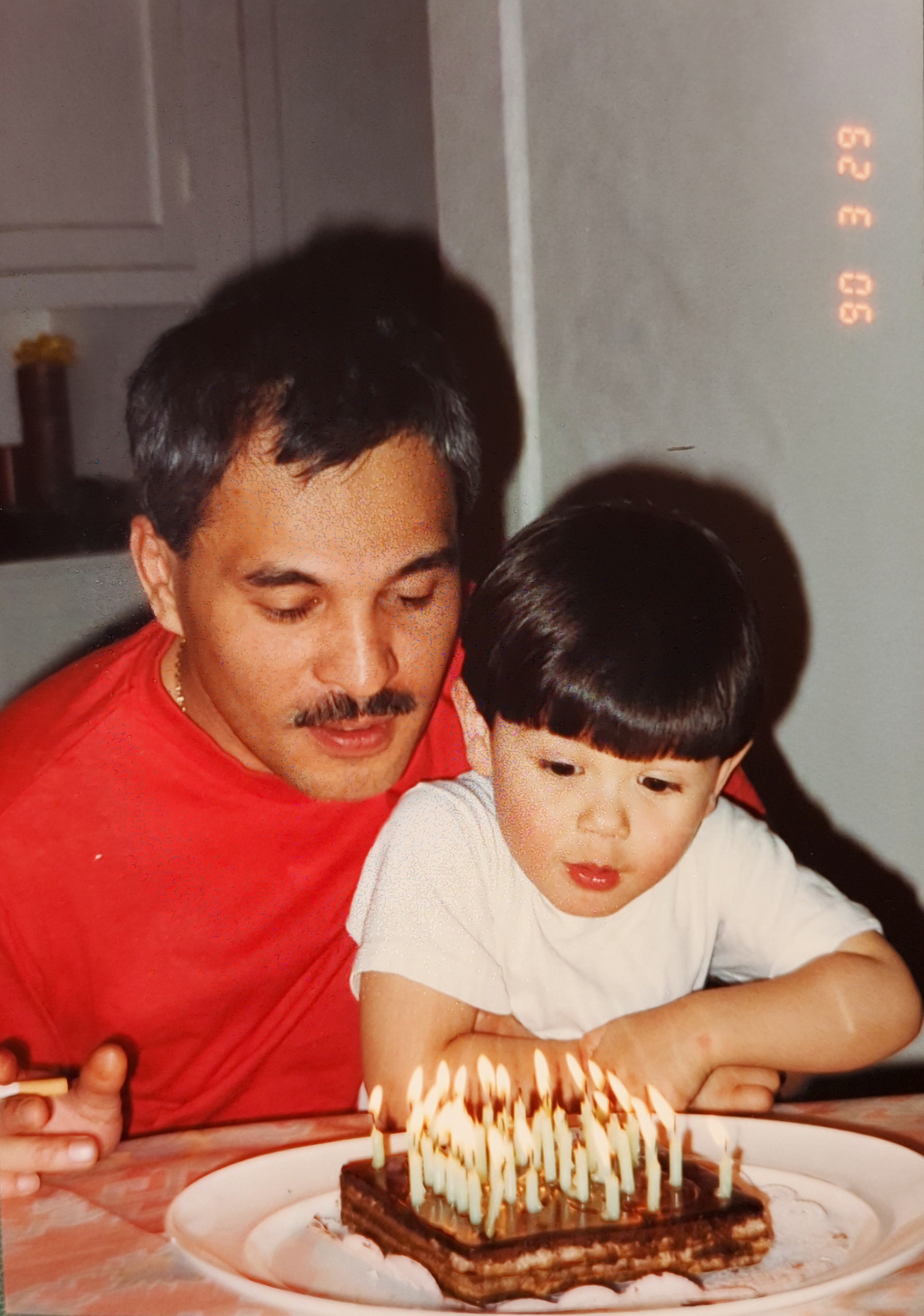 Me and Papa on his 38th birthday. I'm about 3 years old sitting in his lap in front of a birthday cake with many candles on it. I have a bowl cut.